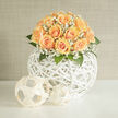 A masterclass in subtlety, this hand-assembled bouquet brings together creamy peach roses and airy white gypsophila in a composition that feels both contemporary and timeless. The roses range from pale, buttery centers to apricot edges, their petals layered in lush spirals that suggest tenderness and the quiet optimism of new beginnings. Tiny clusters of baby's breath weave between the roses like spun sugar, diffusing light and adding an ethereal softness that recalls the veiled mornings beside the Radcliffe Camera. The blooms rest in a sculptural, white-woven spherical vessel-an open rattan orb whose organic latticed lines lift the arrangement, giving it a floating, modern elegance. Green leaves and subtle stems provide structure and a fresh herbal note that refreshes the sweeter floral scent. Sunlight skimming across the scene warms the peach tones and highlights the textural contrast between velvet petals and the crisp, cool weave of the vase. This piece is ideal for weddings, anniversary centrepieces, or a refined gift that speaks of hope and celebration; its presence would sit beautifully on a table in the Covered Market or as an understated focal point in a college dining hall. Crafted with attentive hands and local knowledge, this style of arrangement is available for delivery across central Oxford through Flowers Oxford, reflecting the quiet, artisanal approach our customers expect.
