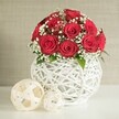 A carefully composed floral still life fills the frame, centred on a dense, rounded bouquet of red roses displayed in a sculptural white vase. The roses, probably around a dozen to fifteen, are in their prime, each bloom unfolding in a tight spiral that reveals layer after layer of velvety crimson petals. Threaded between the roses are generous sprigs of white baby's breath, their tiny blossoms catching the light and introducing an airy, cloud-like feel that softens the strong, romantic red. Occasional flickers of green foliage peek out, just enough to signal fresh stems and to deepen the visual texture. The flowers rise from a distinctive round vase whose entire body is formed from an open lattice of white strands, reminiscent of hand-woven wicker or rattan twigs arranged into a bulbous nest. This textural vase rests on a pale, smooth tabletop, free from distraction, much like a minimalist wedding table set up in an Oxford hotel near the Covered Market or along the Cowley Road. To the left, two decorative spheres lie on the same surface, created from pale, flat strips woven into loose, hollow balls; one is larger, drawing the eye first, while the smaller orb sits slightly behind, creating a subtle play of perspective. The background is a softly blurred wall in a gentle beige or stone hue, its fabric-like texture understated so the arrangement remains the unquestioned star. The lighting is soft and even, with no harsh shadows, enhancing the sense of calm sophistication and making this scene ideal for a romantic wedding table arrangement in Oxford's city centre.