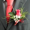 The image offers an intimate view of a wedding buttonhole, secured to the lapel of a charcoal grey suit jacket as though the wearer is about to step into an Oxford college courtyard for photographs. At its heart sits a striking rosebud in deep fuchsia, the petals a rich, velvety pink that spiral inwards, still mostly closed yet full of promise. Encircling this main bloom is a ring of delicate white gypsophila, each tiny flower forming a light, airy cluster that softens the strong colour of the rose and gives the arrangement a romantic, bridal feel. The boutonniere is framed by a thoughtfully arranged mix of greenery: smooth, glossy leaves that provide a solid backdrop and slender, feathery fronds that echo the natural, slightly wild look of foliage from Oxford's riverside paths. At the base, a narrow, bright red satin ribbon is tied into a small, precise bow, its sheen echoing the polished surface of the smooth red silk tie tucked beneath the lapel. The suit appears to be made from a fine wool blend in a dark charcoal tone, its subtle texture visible where the fabric catches the light, and just beyond the tie, a grey dress shirt with a soft pattern adds another layer of refinement. Gentle, even lighting brings out the richness of the rose and the shine of the ribbon without glare, highlighting every curve and contour. The scene feels calm and anticipatory, capturing that quiet moment before a ceremony in Oxford, when every detail-from buttonhole to bow-has been carefully chosen to honour love, tradition, and personal style.