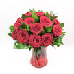 A luxurious bouquet of deep crimson red roses sits at the centre of the image, their velvety petals layered in generous spirals that move from tightly furled hearts to fully opened, cupped blooms. Each rose looks perfectly fresh, as if it has just been conditioned by an experienced florist, with no blemishes on the petals and a rich, saturated ruby tone that immediately evokes romance and devotion. Between the roses, slender stems of dark, glossy foliage - likely Ruscus or a similar filler greenery - weave in and out, the leaves a polished, emerald green that sharpens the contrast and adds depth to the arrangement. The bouquet is gathered into a translucent ruby-red glass vase, whose gently twisted, spiralled surface catches the light and sends soft highlights and shadows dancing across the table. The vase has a slightly flared neck and solid base, giving the whole design a feeling of stability and classic elegance, while the faint outline of the rose stems can be seen through the coloured glass. Everything is set against a pristine white background, so the red and green tones appear especially vivid, almost glowing under bright, even lighting. There are no distractions, just the intense colour and texture of the flowers and leaves. The bouquet feels like it could be waiting on a dining table in an Oxford townhouse near Jericho or Summertown, ready for a candlelit anniversary dinner, a Valentine's surprise, or a heartfelt thank-you. You can almost imagine the soft, sweet fragrance of fresh roses drifting up as you lean in, carrying with it a sense of warmth, passion, and sincere affection.