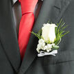 Pinned neatly against the dark charcoal fabric of a formal suit, this classic white rose boutonniere captures the understated elegance often favoured at weddings across Oxford, from intimate gatherings in Jericho to grand celebrations in historic colleges. The central ivory rose is perfectly opened, its petals curving outward in a natural spiral, each one smooth, velvety, and softly luminous against the darker background. Encircling the bloom, sprays of dainty baby's breath cluster like tiny white stars, filling the gaps with airy texture and a sense of tenderness. Slim fronds of fern and gentle loops of glossy green foliage extend outward, framing the rose while suggesting a walk through a leafy corner of Christ Church Meadow. At the base of the arrangement, a crisp white satin bow is tied with careful precision, anchoring the stems and echoing the formality of a freshly pressed shirt and polished cufflinks. The lighting in the image is soft and even, enhancing the contrast between the pale flower and the dark lapel, giving the whole piece a polished yet romantic mood. You can almost imagine the faint, fresh scent of the rose mingling with cool evening air as guests arrive at a city-centre venue, cameras poised. This boutonniere feels suited to a groom, best man, or treasured guest, a quiet but meaningful finishing touch chosen for life's most memorable milestones.