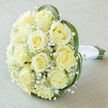 There is a serene, almost dreamlike quality to this bridal bouquet, captured from above so the rounded, opulent dome of blooms reads clearly in soft daylight. About fifteen to twenty pale, creamy-yellow roses crowd together at the centre, their ivory petals unfolding in perfect spirals that catch the light like satin. Interspersed between those lush roses, tiny clusters of baby's breath (Gypsophila) form delicate white clouds that add an ethereal lift and a graceful contrast to the denser rose heads. Long, narrow leaves-resembling bear grass or lily grass-are threaded through the perimeter, their vibrant green arcs suggesting gentle motion as if stirred by the breeze along the Cherwell or in the Botanic Garden. The natural stems peek out at the bottom, thoughtfully bound with a pristine white satin ribbon that has been wrapped and finished with a tight knot to ensure comfort and elegance in the bride's hand. The bouquet sits on a light, subtly textured linen cloth in off-white-taupe tones that complements the soft palette and reinforces the feeling of calm sophistication. The lighting is diffused and soft, emphasizing translucency in the petals and the tender, slightly sweet fragrance that such cream roses typically give-a scent that might mingle with Oxford's spring air near the Covered Market. This arrangement feels like a quiet promise: refined, tender, and perfect for ceremonies, anniversaries, or a refined gift for someone beginning a new chapter.