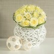 A meticulously crafted floral centerpiece radiates calm elegance, built around a rounded cluster of light yellow roses that fill the frame with their soft, creamy glow. Each rose is at its peak, petals unfurling in generous spirals so that one bloom flows seamlessly into the next, creating a plush, cushion-like surface. Interwoven between the roses, tiny white florets of baby's breath lend a veil of delicate texture, lightening the bouquet and giving it a romantic, wedding-ready character. At the bottom, a narrow band of fresh green foliage is wrapped with precision, delineating the edge of the flowers and drawing the eye down to the sculptural white vase. This vase resembles a modern piece of basketry, its slender, white strands crossing and looping to form an airy, open structure that allows glimpses of shadow and reflected light. Resting on the glossy white tabletop beside it are two decorative spheres made from pale, ribbon-like strips, their woven pattern echoing the vase so the whole composition feels thoughtfully curated. In the background, a soft, neutral beige backdrop blurs gently, much like the warm stone façades found throughout central Oxford, from Broad Street to the lanes by the Sheldonian. The overall effect is sophisticated yet inviting, perfectly suited to a wedding table in an Oxford college hall, a chic restaurant reception on the High Street, or a romantic engagement dinner at home. The image communicates artistry, balance, and an atmosphere of tender celebration, as if ready for same-day delivery to set the scene for a special occasion.