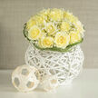 Creamy yellow roses form a compact, dome-shaped bouquet here, each bloom edged with ruffled, satiny petals that open like small suns. Interlaced among them, clouds of white gypsophila float and breathe, softening the silhouette with tiny stars of texture. The flowers rest inside a striking white wicker orb-an airy, modern vessel whose crisp weave reads as both container and sculpture-while a few curling blades of greenery loop around the top, offering gentle movement and framing the roses with an understated rhythm. Nearby, simple woven spheres add a contemporary playful beat that complements the basket's geometry and invites the eye to travel around the arrangement. Soft, natural light from a northwest window-reminiscent of the calm mornings along Oxford's High Street-lends the roses a warm glow, and there is a subtle fragrance: a clean rose perfume with a faint hint of summer meadow and freshly turned earth beneath. This bouquet feels especially suited to quiet celebrations-a thoughtful gift for a new academic appointment at the university, a serene centrepiece for a small gathering in Summertown, or a warm expression of encouragement delivered to a friend. Crafted with careful balance and an eye for texture, it reflects local sensibility and floral expertise. Flowers Oxford offers similar compositions for local delivery across the city, bringing this refined, tactile harmony to doorsteps from Cowley to the Covered Market.