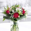 A lush red and white bouquet stands proudly in a clear cylindrical glass vase, its neatly trimmed green stems visible through the water and giving a sense of absolute freshness, like something just arranged in a boutique off Oxford's High Street. Deep red roses, their velvety petals slowly unfurling, create a strong romantic heart to the arrangement, echoed by a large ruffled crimson carnation at the front whose intricate, layered petals add rich texture and drama. Around these bold blooms, clusters of white and creamy flowers provide contrast and softness: white alstroemeria with slender petals marked by delicate speckles, tight white lily buds promising future blossoms, and airy clouds of tiny gypsophila that look like a gentle sprinkling of snow. Glossy dark green tropical leaves are carefully curled and wrapped around the base, forming an elegant collar that cradles the flowers, while varied foliage in eucalyptus-like tones and pointed green leaves fill out the shape and add depth. The bouquet rests on a spotless white surface, possibly a table in a bright Oxford townhouse near Christ Church Meadow, with a softly blurred, light-filled background suggesting a sunny window. Even, natural-style lighting catches each petal and leaf, highlighting the bouquet's celebratory reds, peaceful whites, and abundant greens. The whole composition feels poised yet warm, ideal as a refined centrepiece for an anniversary dinner, a thoughtful Mother's Day delivery across Oxford, or simply to bring heartfelt affection and a touch of classic romance into a home or office.