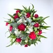 A richly detailed, top-down photograph of a hand-tied bouquet shows a full, circular arrangement of red and white flowers resting against a spotless white background, the colours glowing as if lit by soft natural daylight over Oxford's High Street. Deep crimson roses form the heart of the design, some still in tight, velvety buds, others just beginning to unfurl their layered petals, suggesting quiet romance and enduring love. Nestled between them are ruffled carnations in vivid fuchsia and ruby red, their frilled, cushiony blooms giving the bouquet a festive, almost celebratory energy, like a gathering after a ceremony at the Sheldonian Theatre. Clusters of small white spray chrysanthemums, with daisy-like faces and neat yellow-green centres, add a fresh brightness, echoing the clean backdrop and lifting the whole palette. Tiny white gypsophila, or baby's breath, is scattered generously throughout, its cloud of star-like florets softening the spaces between the larger flowers and creating a misty, delicate halo. Surrounding and weaving through the blooms is lush greenery: broad, pointed dark green leaves from lilies or alstroemeria anchor the design, while silvery-green eucalyptus leaves, rounded and slightly matte, curl gently at the edges, adding a contemporary twist and hinting at a cool, herbal scent. The bouquet appears dense and luxurious, crafted with care and balance, ideal for sending a message of love, gratitude, or heartfelt congratulations. It feels like the kind of arrangement a local Oxford resident might send across Summertown or Jericho to mark an anniversary, a birthday, or to brighten a quiet townhouse with a burst of colour and life.