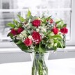 A luxurious bouquet of red roses, ruffled red carnations, white alstroemeria, and clouds of tiny white gypsophila stands in a clear glass vase on a bright white tabletop, bathed in soft natural light from a large window, like you might find in a calm Oxford townhouse near Christ Church Meadow. The roses are a rich crimson, some fully open with velvety petals spiralling outward, others still in tight, elegant buds promising more colour in the days ahead. Nestled between them, the carnations echo the same deep red hue, their frilled, multi-layered petals adding fullness and a comforting, traditional charm. Clusters of white alstroemeria, many still in bud with faint green tips and a few partially open showing their graceful, trumpet-shaped blooms, provide a gentle contrast to the bold reds. Sprays of baby's breath weave through the arrangement like a fine mist, tiny white flowers forming a soft, cloudlike halo that lightens the entire design. Lush greenery anchors the bouquet: broad, glossy deep green leaves form a collar at the base, while slimmer, lighter green stems and foliage thread upwards, creating movement and texture. The stems are clearly visible through the cylindrical vase, neatly cut and submerged in fresh water, reinforcing the sense of just-arrived freshness typical of same-day local delivery across Oxford. Behind the bouquet, the view through the window is softly blurred and almost monochrome, allowing the reds, whites, and greens to stand out with striking clarity. The overall mood is warm and celebratory, perfect for marking an anniversary on the Cowley Road, a romantic gesture in Jericho, or simply brightening a quiet Corner of a Summertown home with a touch of classic floral elegance.
