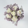 High above a small, hand-tied bouquet, the camera looks straight down onto a perfect circle of flowers: six creamy white roses resting at the heart of a cloud of lilac and lavender statice. Each rose is tightly formed yet gently opening, with layers of smooth, velvety petals spiralling toward the centre, their ivory tones carrying the faintest blush of green at the base of the sepals. The roses appear almost luminous against the haze of tiny purple blossoms that surround them, the statice creating a soft, feathery texture like a mist rolling over Port Meadow on an early Oxford morning. The filler flowers are densely packed but airy, filling every gap so that no stem or mechanics are visible, only a continuous cushion of mauve and lavender shades that frame the pristine blooms. The bouquet is placed on a pale, off-white surface, similar to a clean tabletop in a stylish Jericho townhouse, the neutral background allowing the flowers to take full focus. Soft, even overhead lighting adds a calm atmosphere, with no harsh shadows, only gentle shading that brings out the petals' delicate curves and the statice's fine details. There is no visible vase or ribbon, just the clear sense of a compact, hand-held arrangement designed for intimate gifting-a tender posy suitable for a quiet anniversary, a thoughtful condolence, or a peaceful bedside arrangement. The overall feeling is one of serenity and refinement, a classic white-and-lilac bouquet that would look perfectly at home delivered to an Oxford college room, a small wedding gathering in Summertown, or a restful corner of a home in Headington, radiating purity, comfort, and understated elegance.