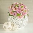Beneath the soft, inviting glow of natural afternoon light in Oxford, a beautiful handcrafted floral composition becomes the focal point of a meticulously set table. The centre of attention is a lush, rounded bouquet of pastel-hued blooms-soft blush pink roses mingle with lavender roses, their velvety petals radiating serenity and freshness. Each is complemented by trumpet-shaped pink alstroemeria, tracing the unique striped and spotted patterns that add a bespoke artistic touch, not unlike the diversity seen at local Oxford flower markets. Bursts of vivid lime green button chrysanthemums weave through the arrangement, their playful pompom forms catching the eye amidst interspersed sprays of airy white baby's breath. Glossy, deep green leaves emerge between blooms, rooting the design in the vivid palette of the surrounding countryside. All are presented in a statement vase, a white, open-lattice vessel crafted from slender, intertwining strips-its unique basket-like weave and gently bulbous shape imparting a sense of rustic charm, reminiscent of the historic crafts found in Oxfordshire villages. At the left, resting on the crisp white table, two decorative spheres of different sizes echo the vase's design, fashioned from pale woven strips and positioned for balance and fluidity. The neutral, textured beige backdrop enhances the delicate colour story and tactile craftsmanship, evoking a sense of artisanal warmth and welcoming sophistication. Whether as a bespoke centerpiece for an intimate Bodleian Library celebration or a thoughtful gift to brighten a Jericho townhouse, this arrangement embodies the artistry and heartfelt attention to detail for which Oxford florists are renowned.