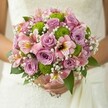 A bridal bouquet, overflowing with romance and freshness, rests in the gentle grasp of a bride against her lace-adorned Oxford wedding gown. Each element sings of new beginnings and heartfelt celebration: dusty lilac and soft pink roses anchor the design, their unfurling petals reminiscent of historic blooms found in the walled gardens along High Street. Vibrant green button chrysanthemums add a burst of lively energy, their playful shapes echoing youthful laughter and the verdant lawns of Oxford colleges. Slender alstroemeria lilies, their blush petals painted with streaks of maroon and sunlit yellow, weave an air of subtle exoticism through the arrangement. Sprays of wispy white baby's breath drift between the fuller blossoms, creating a delicate backdrop that shimmers in the morning light, reminiscent of early mist over the Cherwell. Lush green foliage surrounds the bouquet, grounding the colourful display with its deep emerald hue. The entire composition feels luminous, serenely joyful, and perfectly suited for an Oxford celebration-be it a stately gathering beneath dreaming spires or an intimate riverside ceremony. This bouquet speaks of expert hands, fresh flowers gathered at dawn, and the enduring beauty that only a local Oxford florist can bring to such a meaningful occasion.