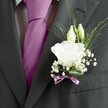 A sharply focused close-up of a groom's charcoal grey suit jacket shows a meticulously crafted white lisianthus buttonhole resting against the lapel, the kind of refined detail you might notice at a wedding in an Oxford college chapel. The wool suit has a subtle textured weave, its crisply pressed lapel framing a deep magenta silk tie knotted perfectly at the centre of the image. Beneath the tie, a pale grey and white patterned dress shirt peeks through, adding quiet structure. Pinned to the left lapel is a handcrafted boutonnière: one fully opened white lisianthus bloom with softly ruffled petals, edged like fine tissue, sits at the heart of the arrangement. Two slender ivory buds hover just above, still closed and full of promise, surrounded by a light cloud of tiny white gypsophila blossoms that soften the overall shape. Fine strands of airy greenery arc outward, lending movement and a fresh, garden-picked feel, as if gathered from a morning stroll along the Cherwell. At the base, a narrow mauve ribbon, carefully wrapped and tied into a neat bow, echoes the tone of the tie and anchors the stems tidily against the jacket. The lighting is even and gentle, highlighting texture rather than glare, and the composition draws the eye to the harmony between tailored fabric and delicate petals. The image conveys calm anticipation and quiet romance, perfectly suited to a groom or groomsman preparing for a ceremony in central Oxford, where every small floral detail is thoughtfully considered and designed to complement formal attire without overwhelming it.