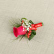 This image shows a thoughtfully crafted boutonniere centred on a single hot-pink fuchsia rose, its petals dense, ruffled, and elegantly unfurling as though caught at the height of its beauty. Tiny white gypsophila, often called baby's breath, sits in soft, cloud-like clusters around the rose, each miniature blossom catching the light and adding a veil of softness to the composition. Nestled between the blooms are red hypericum berries, rounded and lightly lustrous, bringing an extra note of celebration with their lively, berry-red tones. Encircling everything, polished green leaves are folded and curled in smooth arcs, including broad, ribbon-like aspidistra that overlaps in graceful loops, and slender blades of decorative grass that trail and flick out, lending a gentle sense of movement. The stems are gathered and wrapped in a rich red ribbon, spiralling tidily downwards to create a secure and refined handle that would sit comfortably against the lapel of a suit or jacket. The boutonniere lies on a softly woven, neutral background, perhaps a linen or cotton fabric, its subtle texture helping the saturated colours stand out as they might against a shirtfront at a ceremony in central Oxford, near the Bodleian Library or along the High Street. In mood, the piece feels romantic yet modern, with its clean lines and clear colour contrasts. You can imagine the light scent of the rose mingling with the freshness of the greenery, and the cool, slightly waxy touch of the foliage under the fingertips. It carries a quiet promise of celebration, perfect for weddings, proms, or intimate city gatherings by the river in Oxford.