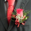 A close-up, high-angle photograph reveals an impeccably styled buttonhole on the left lapel of a charcoal grey suit, the kind you might see on a groom stepping out of a classic Oxford townhouse near Jericho for his wedding. At the heart of the arrangement sits a single deep red rosebud, petals tightly furled yet plush, catching the soft indoor light with a velvety sheen. Surrounding the rose, tiny white filler blooms, similar to baby's breath, float like a delicate mist, their starry shapes softening the intensity of the crimson. Nestled among them are small, glossy red berries, round and polished, echoing the rose's rich tone and adding a subtle, tactile texture. Slim, grass-like blades of greenery fan out behind the flowers, while broader, darker green leaves curl protectively at the base, giving the design structure and depth, as though it had just been gathered from a garden walk along the River Thames by Folly Bridge. A neat red satin ribbon is wrapped and tied into a precise little bow at the stem, its sheen perfectly in tune with the hue of the rose and berries. Beneath the lapel, a vivid red tie, likely silk, is knotted crisply against a finely patterned shirt in pale grey and white, adding a refined, almost scholarly elegance that suits the city's academic character. The overall impression is one of carefully curated formality and romance, an image that speaks of thoughtful boutonnière selection and colour coordination for weddings, proms, or black-tie celebrations across Oxford's historic colleges and elegant venues.