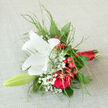 This hand-tied bouquet appears as a small, vivid story in flowers, laid gently against a pale cloth as if ready to be carried down an aisle in Oxford's old city centre. A single white lily takes pride of place, its petals elegantly flared, smooth and luminous with a creamy glow that suggests purity, clarity, and a quiet sense of optimism. Nestled close beside it, deep red roses unfold in layered spirals, their velvety texture and rich tone evoking enduring love, much like the timeless architecture along Broad Street or the tranquil paths through University Parks. Clusters of bright red berries peek out between the blooms, shiny and round, lending a joyful, almost festive spark, while soft sprays of gypsophila weave in and out like delicate lace, adding lightness and a touch of innocence. Wispy green foliage arches around the flowers, feathery and gently tousled, giving the composition a natural movement, as though a breeze from the Cherwell had just brushed through it. The stems are carefully aligned and bound in a slim band of scarlet satin ribbon, its smooth finish and subtle sheen suggesting thoughtful craftsmanship and attention to detail by an experienced Oxford florist. The arrangement carries an atmosphere that hovers between celebration and gentle reflection, equally at home in a wedding celebration, an anniversary surprise in Summertown, or a tender gesture of support during an important life moment. You can almost imagine the mix of scents-the lily's soft perfume, the faint sweetness of the roses-combining into a fragrance that lingers like a kind memory, comforting and sincere.