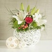 A romantic and refined floral arrangement is presented as a low, abundant centerpiece, ideal for wedding tables in Oxford's charming hotels and college halls. It rests in a distinctive round white vase, whose open-weave design-formed from intertwining white strands-recalls a modern interpretation of a woven basket, with small pockets of light and shadow that add depth to the composition. From this textured vessel, a garden of white, red, and green seems to spill outward. Several white lilies dominate the scene, their long stems rising gracefully and their blooms at different stages of opening: some fully unfurled with broad, creamy petals speckled softly near the centre, others still sheathed in smooth green buds that suggest fresh flowers yet to come. Nestled close to these lilies is a single striking red rose, its velvety petals tightly layered into a lush spiral, giving the arrangement a focal point of passionate colour that feels perfect for toasts and speeches at an Oxford wedding reception near the Bodleian. Bright green, ball-shaped blossoms-likely green dianthus or pompon chrysanthemums-pepper the design with an almost moss-like texture that feels playful and contemporary. Wisps of white baby's breath weave delicately through the bouquet, softening the transitions between colours, while small clusters of red berries add another layer of interest and a subtle festive note. A mix of greenery, from feathery fronds to broader dark leaves, fills out the base and spills gently over the rim of the vase, giving the piece a natural, slightly untamed character. Beside the main arrangement, two light, woven decorative spheres echo the vase's texture, tying the scene together against the understated neutral background, like a thoughtfully styled wedding table awaiting guests somewhere between the city centre and Summertown.