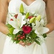 This quintessential Oxford bridal bouquet evokes the atmosphere of a secret university garden at dawn: classic, fresh, and effortlessly elegant. Large white lilies, their petals unfurling with confidence, frame the composition against the bride's lace-trimmed gown-a nod to Oxford's storied craftsmanship. Tucked amongst the lilies, crimson roses glow with romantic intensity, their velvet spirals promising devotion that endures far beyond wedding day festivities. Clusters of red hypericum berries peep out playfully, infusing radiant bursts of colour akin to those seen at local florists' stalls on the High Street. Wispy baby's breath weaves throughout, casting an airy, dreamlike spell. Deep green ferns and button-like mossy spheres create a grounding harmony at the bouquet's base, conjuring the lush mystery of Christ Church Meadow after gentle rain. The arrangement is a tactile joy-silken petals, cool foliage, and buoyant berries all held in the bride's gentle grasp, the emotion of the moment captured in soft, golden light. Created for a wedding woven into Oxford's rich tapestry-from the colleges to the quads-this bouquet tells a story of love, commitment, and fresh beginnings, lovingly arranged to linger in the heart long after the vows are spoken.