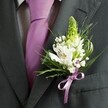 A high-angle close-up captures the upper torso of a groom or groomsman dressed in a finely tailored charcoal grey suit, the dark fabric forming a smooth, structured backdrop for a wedding buttonhole. The lapel is sharply cut and sits over a pale dress shirt patterned with a subtle grid, while a sleek lavender tie, neatly knotted, draws the eye down the centre of the chest. Pinned precisely to the left lapel is a detailed boutonniere: a tall, pale green spire of tightly closed buds rises at the top, giving gentle height and a sense of anticipation, while a cluster of white alstroemeria blooms opens below, each petal etched with fine maroon streaks and soft golden centres. Around them, airy sprays of white gypsophila, or baby's breath, create a cloud-like halo, their tiny florets catching the light and softening the composition. Slender, grass-like blades of greenery curve outward, interwoven with slightly broader, glossy dark green leaves that frame the flowers and contrast with the suit's charcoal tone. At the base, a satin ribbon in lilac-lavender is tied into a small, perfectly formed bow that echoes the colour of the tie, adding a gentle touch of romance. The lighting feels natural and calm, perhaps like a quiet morning in Oxford before guests gather at a city-centre venue or along the cobbled streets near the High. The overall mood is one of poised elegance and fresh celebration, conveying the quiet emotion of a wedding day just about to begin, with every detail, down to this single lapel flower, thoughtfully curated and ready for photographs and heartfelt vows.