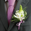 This image offers a detailed, intimate view of a gentleman's formal attire, as if you were standing just in front of him on an Oxford church step moments before the ceremony begins. The suit jacket is a deep charcoal grey, finely tailored with a clean lapel edge and subtle seams that frame the boutonniere. Beneath sits a pale, patterned shirt, likely white with a discreet dark check, providing a soft, almost woven-looking backdrop. A solid lavender tie falls smoothly down the centre, its satin surface catching soft light like reflections off the River Thames by Port Meadow, bringing a calm, modern note to the look. Pinned to the left lapel, the boutonniere showcases white alstroemeria blooms, their petals marked with delicate reddish-brown brushstrokes and small yellow centres, each flower crisp and fresh, as though arranged that morning by a local florist on the High Street. Standing upright in the middle is a slender, conical spike of lime-green buds, adding height, texture, and a hint of budding fragrance. Around it, clusters of white baby's breath drift like tiny clouds, interlaced with fine, grass-like blades and feathery greenery that soften the outline. Broader, glossy green leaves cradle the arrangement, giving structure and a lush, tactile depth you can almost feel beneath your fingers. At the base, a lilac ribbon bow, perfectly tuned to the tie, gathers the stems into a neat, polished finish. The overall impression is of thoughtful coordination and quiet celebration, an elegant corsage-style accent suited to weddings, proms, or black-tie events anywhere from Jericho to Headington.
