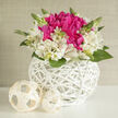 A round white wicker basket holds a lush, carefully composed bouquet that seems to glow against its minimalist setting. At its heart, rich fuchsia blossoms resemble exuberant peonies, their many ruffled petals layered like luxurious fabric, creating a focal point full of depth and romance. Surrounding them, creamy white alstroemeria form a soft, embracing collar, each slender petal decorated with delicate brushstrokes of pink and fine speckles that add quiet intrigue. Fresh green leaves, tender stems, and tiny buds weave in and out of the arrangement, introducing a feeling of new growth, like the first shoots appearing in an Oxford allotment after a gentle rain. The basket itself is a piece of craftsmanship, woven from crisp white strips into a rounded vessel that feels both contemporary and classic-something you might find gracing a wedding breakfast table in one of Oxford's dreaming spires or boutique hotels. At the base, two pale cream orbs with a similar woven texture rest on a white surface, echoing the basket and anchoring the image with calm balance. Soft, diffused light spills over the flowers, highlighting the contrast between the vivid pink centre and the tranquil ivory ring around it, and creating small pools of shadow that add depth without drama. The mood is romantic and refined yet welcoming, ideal for bridal bouquets displayed at an Oxford Town Hall ceremony, centrepieces for a Cowley Road celebration, or a special delivery to mark an engagement. The whole design feels thoughtfully curated, as if arranged by a florist who knows the rhythm of local weddings and the quiet joy of giving flowers in Oxford.
