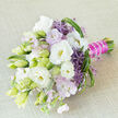 A sophisticated hand-tied bouquet lies diagonally across a subtly textured light beige fabric, its colours balanced with the same care a florist at Flowers Oxford might give to a wedding order near Summertown. The arrangement centres on lush white lisianthus, their petals layered into soft ruffles that catch the light, with some blooms fully open to reveal creamy yellow throats, while others remain as elegant, tapered buds in pale green and ivory. Threaded among them are delicate sprays of lavender-pink blossoms, fine and feathery, which add a pastel haze and soften the overall silhouette. A handful of vivid purple allium florets, shaped like tiny spiky stars, rise up as sculptural accents, offering a lively contrast to the smooth, rounded forms of the lisianthus. Around the flowers, varying shades of fresh green foliage create depth and texture: long, slender leaves, occasional curled strands, and lighter, spiky tufts give the bouquet a sense of movement, as if stirred by a gentle breeze drifting across an Oxfordshire meadow. The stems are neatly trimmed and bound in a bold fuchsia satin ribbon, wrapped with precision so that the handle appears smooth and luxurious in the hand. Along the length of the ribbon runs a refined strand of small white pearls, lending a note of classic elegance that would suit a ceremony in one of Oxford's historic colleges or a stylish celebration in Headington. The imagined scent is subtle-fresh, slightly sweet, and calming-making this bouquet ideal for weddings, anniversaries, or a meaningful same-day delivery across Oxford that speaks of romance, modern elegance, and carefully curated detail.