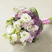 A carefully hand-tied bouquet lies gracefully on a light beige, linen-like cloth, the neutral backdrop allowing the flowers' gentle colours to glow as if lit by soft morning light drifting over Oxford's High Street. At the heart of the arrangement, white lisianthus blooms unfurl in layered whorls of petals, some fully open to reveal pale yellow-green centres, others still curled in tight buds that promise to open later in the day. Around these central blooms, clusters of soft pink and light mauve stock flowers form a plush, cloudlike surround, their many tiny, ruffled petals adding a sense of volume and romance. Striking clusters of purple, star-shaped filler flowers-much like ornamental allium-emerge between the pastel tones, their spiky textures and rich lavender hue bringing an intriguing contrast that feels both modern and whimsical. Long, slender strands of deep green grass-like foliage run through the composition, looping and arching, particularly along the right side, creating a feeling of movement reminiscent of the breeze in University Parks. The stems are gathered neatly and bound with a vivid fuchsia satin ribbon, the fabric smooth and lustrous, providing a bold note of colour that accentuates the feminine softness of the blooms. A fine strand of small, round white pearls is wrapped over the ribbon, catching the light in tiny reflections, like raindrops on college cloisters after a spring shower. This bouquet evokes calm celebration and gentle affection, ideal for a bride stepping out of an Oxford town hall ceremony, a special anniversary dinner in Summertown, or any occasion where subtle luxury and heartfelt beauty are cherished.