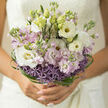 A hand-tied bouquet rests in the frame like a whispered promise, its colours and textures echoing the soft, misty mornings along the Oxford Canal. At its heart, creamy white lisianthus unfurl in generous, rose-like whorls, each petal edged with a faint, translucent sheen that catches the daylight. Interwoven with them, slender stems of stock in pale lilac and off-white rise gently, their clustered blossoms adding height and a cloud-like softness to the arrangement. At the base, a circular band of allium blooms in dusky lavender and warm purple provides both structure and intrigue, every tiny floret forming a miniature star that draws the eye downward. Encircling everything, thin strands of deep green bear grass spiral and loop with deliberate artistry, creating a modern halo that frames the bouquet without weighing it down. The overall impression is light yet composed-airy at the top, more architectural at the base-like a carefully composed photograph taken near Oxford's Radcliffe Camera in late afternoon sun. The colour palette remains refined: ivory, pastel lilac, touches of blush, and vibrant green foliage combining into a gentle, romantic spectrum. You might imagine this bouquet being carried through narrow lanes off the High Street on the way to a quiet civil ceremony, the cool, faintly sweet scent of the lisianthus and stocks mingling with the crisp city air. It conveys a sense of poised expectation and thoughtful elegance, a floral reflection of those cherished, hope-filled moments that mark the start of a shared life in this historic city.