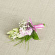 Resting on a softly woven, neutral cloth, this petite boutonnière appears like a carefully composed still life, the kind a local florist in Oxford might prepare on a calm morning before deliveries head out towards Jericho or Headington. At its centre, creamy buds-likely freesia or similar delicate blooms-are only partially open, their petals smooth and almost translucent, giving the impression of fragile porcelain. They sit alongside blush-pink flowers that unfold in gentle layers, each petal slightly curved and silky, offering a tender contrast of tone and texture. Above and between them, fine sprays of baby's breath float like tiny white clouds, softening the outline of the arrangement and bringing a sense of light, airy romance. Slim, blade-like strands of greenery weave through the design, echoing the loose grasses and hedgerows seen around Oxford's quieter lanes, while broader leaves curl neatly at the base, providing structure and a natural, verdant frame. The florist has bound the stems tightly with a pale rose-pink satin ribbon, wound with precision and finished in a small, perfectly tied bow that suggests care and craftsmanship. Under soft, diffused lighting, the interplay of ivory, blush, and fresh green looks calm and harmonious, as if captured just before being pinned to a lapel for a wedding at a historic Oxford college or a celebration in a converted barn on the city's edge. The imagined fragrance is subtle yet evocative-fresh, slightly sweet, and clean-giving this boutonnière an intimate, personal quality, like a gentle floral whisper that accompanies the wearer through a day of heartfelt moments.