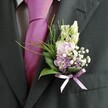 A detailed close-up reveals the upper body of a man dressed for a special occasion, his charcoal grey suit jacket filling most of the frame against a softly blurred background. The fabric of the jacket has a fine woven texture that catches the light along the lapel, while a pale dress shirt with a subtle grid pattern peeks out neatly at the collar and cuffs. At the centre of the image a bold, smooth silk tie in vivid magenta, almost fuchsia, runs straight down the chest, its sheen suggesting careful pressing moments before the ceremony. Pinned to the left lapel, a carefully crafted boutonnière becomes the real focal point. Creamy, unopened buds-reminiscent of slender snapdragons-stand upright at the top, their pale ivory tones gently transitioning into clusters of soft pink and light lavender flowers below, similar to sweet peas or fragrant stock. Tiny clouds of white gypsophila are threaded between the blooms, their airy dots of colour adding a mist-like softness, while a few deeper purple florets provide quiet depth. At the base, glossy dark green leaves cradle the stems, anchored by brighter, feathery sprigs of foliage that arc lightly outward, echoing the natural movement of a riverside breeze along the Thames in Oxford. A delicate lavender satin ribbon is tied into a neat bow, hugging the stems and harmonising with the pastel tones above. Bathed in gentle daylight, the flowers appear freshly arranged, ready for a wedding at an Oxford college chapel or an intimate ceremony near the Covered Market, capturing both elegance and a playful joy in the groom's or guest's attire.