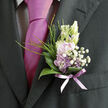 Nestled against the dark, finely woven fabric of a formal suit, this boutonnière offers a tender contrast of colour and texture, the kind of detailed work a local Oxford florist would prepare for a ceremony in Jericho or a celebration in Summertown. Soft lilac flowers form the focal point, their petals gently cupped, with a pastel, almost powdery quality that suggests newly opened blooms. Surrounding them, delicate sprays of white baby's breath create a halo of tiny, star-like blossoms, filling the gaps with a dreamy, airy lightness. A collar of polished green garden leaves curves protectively around the arrangement, their surfaces smooth and reflective, while slender strands of grass add an upward movement that makes the design feel alive and breathing. At the base, a sheer lavender ribbon wraps the stems with a careful, tidy flourish, its subtle shimmer mirroring the tones of the tie beneath and tying the whole ensemble together. The lighting catches each element softly, highlighting the interplay between the cool lilac, bright white, and deep green, so that the boutonnière appears fresh, dewy, and just-arranged. One can almost sense the fresh garden scent, a mix of leafy greenness and gentle floral sweetness, like walking through University Parks on a mild spring morning. Designed on a miniature, intimate scale, this piece is ideal for a groom, best man, or special guest, carrying a quiet message of romance, care, and the craftsmanship that transforms a handful of small flowers into something memorable and deeply personal.