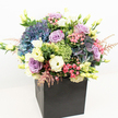 A visually stunning and vibrant floral arrangement bursts forth from a sleek matte black box, its shape and colours expertly balanced to capture the essence of Oxford's lively character. Large, full hydrangeas in shades of indigo and powder blue set a plush foundation, their billowing presence immediately drawing the eye, like a garden in full bloom beside the city's historic colleges. Interspersed with them are softly unfurling roses in lavender and gentle blush, their fragrant petals adding a romantic note, while creamy white eustoma and delicate yellow lisianthus evoke a timeless sense of elegance. Sprigs of textured lime green fillers, such as Green Trick dianthus, add an energetic, playful vibe, while airy pale pink astilbe mimics the gentle movement of a summer breeze along Port Meadow. Accent foliage in a variety of greens-from glossy deep leaves to bright tendrils-emboldens the arrangement and brings it to life. Set against a pristine white background, each bloom practically glows under even lighting, showcasing their freshness and vivid hues. The black gift box grounds the entire composition, offering a sleek, contemporary frame that highlights the arrangement's lush abundance. Thoughtfully designed for all sides to impress, this bouquet embodies sophistication and joyous celebration, making it a perfect choice for luxe gifts, special events, or a memorable delivery anywhere in Oxford's vibrant community.