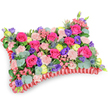 Seen from a gentle, slightly elevated angle against a crisp white backdrop, this pillow-shaped floral tribute is a compact tapestry of colour and texture designed to comfort. The top surface is densely packed with velvety hot pink roses-some tightly budded, others just beginning to unfurl-nestled alongside clusters of pale, shell-pink spray carnations that bring a soft, ruffled surface to the composition. Bright lemon-yellow ranunculus punctuate the pinks with sunny, buttercup-like faces, while deep purple lisianthus (eustoma) create bell-shaped pockets of shadow and contrast. Scattered through the massed blooms are silvery-blue Eryngium thistles, their spiky, architectural forms giving a quiet, wild edge that breaks the otherwise plush textures. Rounded, silvery-green eucalyptus leaves and darker green filler foliage weave through the flowers, adding cool tones and a calming scent that catches the light and lifts the arrangement's depth. The pillow is framed by a rich, deep-red pleated satin ribbon, ruffled around the perimeter to define the classic cushion shape and lend a discreet elegance. Soft morning light in the photograph reveals the subtle sheen of petals and the satiny border without harsh shadows, emphasising the care in placement and symmetry. Traditionally chosen as a sympathetic memorial cushion, this piece reads as a celebration of life-warm, tender and reassuring rather than somber-offering a fragrant, visual embrace for funeral services or graveside remembrance in Oxford, from the Covered Market to the quieter lanes near the Botanic Garden.