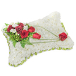 A solemn, carefully crafted rectangular sympathy pillow made entirely from hundreds of small, pristine white chrysanthemum blooms sits against a soft white background. The densely packed chrysanthemums create a plush, cloud-like surface that reads as both tactile warmth and visual calm, the petals forming a velvety field that suggests peace and rest. Around the edge a thin band of light green satin or wired ribbon peeks through the flowers, giving the pillow a gentle, finished frame. On the upper left corner an elegant cluster of deep red roses and rich burgundy carnations forms the focal point; the roses range from tight buds to open, velvety blooms while the carnations' ruffled edges add depth and shadow. Interspersed among them are small white freesia or spray blooms whose delicate shapes lift the composition and soften the strong reds. Lush green foliage-feathery fern fronds and glossy leaves, perhaps with sprigs of eucalyptus-backs the spray, and slender arcs of bright green bear-grass or similar foliage sweep diagonally from that top cluster toward the lower right. This graceful green arc culminates in a single, perfectly formed red rose resting at the pillow's bottom-right, as if gently carried across the white field. The overall scent is quietly floral and clean; the arrangement reads as an expression of lasting love and respect. In Oxford this traditional memorial cushion is often chosen for services at local churches or quiet tributes around Radcliffe Square, offering a dignified, comforting presence for farewell and remembrance.