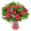 A sumptuous, rounded bouquet of a dozen or more deep crimson roses sits like a jewel in a tall, translucent ruby-red glass vase. Each rose is fully open, its velvety petals spiraling from a tight centre into lush, sensuous layers that catch the soft, even studio light and reveal subtle wine-dark shadows along the petal edges. Interwoven between the blooms is a generous mass of dark green foliage: flat, glossy leaves-reminiscent of salal or Ruscus-form a dense verdant backdrop while broader, emerald Aspidistra leaves have been meticulously curled and rolled into sculptural loops and spirals. Those architectural green forms arc around the rose heads with an almost graphic precision, their glossy surfaces reflecting the same warm highlights as the red glass below. The vase itself is slender and elegant, its faint twisted or ribbed texture creating vertical highlights that visually echo the roses' stems and add a modern rhythm to the composition. Set against a clean white background, the intense crimson and rich greens pop with theatre and clarity, isolating the arrangement like a single, heartfelt statement. There is an implied scent-classic, slightly fruity rose perfume-that seems to hover above the tightly clustered blooms, and the overall effect reads as both romantic and refined. Imagined for anniversaries, Valentine's Day, or an intimate celebration, this arrangement carries a quiet luxury and hand-crafted confidence; it's the sort of bouquet a local florist might prepare for a table in a Georgian townhouse near the High Street or as a romantic gesture sent across town to Jericho or Summertown.