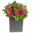This elegantly modern arrangement places rich, velvety red roses at its heart, each bloom opening in tight, generous spirals of deep crimson that read as both tender and dramatic. Broad, glossy green leaves are skillfully curled and woven around the roses, their looping edges and overlapping planes creating a rhythmic texture that leads the eye in a gentle circulation through the bouquet. The flowers sit in a crisp, matte black cube whose square geometry grounds the composition and gives the design a contemporary, urban presence; the contrast between the matte vessel and the luminous petals heightens the visual drama.

Bright, even lighting picks out the sheen on each leaf and the satiny highlights along the rose petals, lending the scene the feel of a freshly opened gift placed on a mantel or a modern dining table. Run a fingertip mentally along the surfaces: the petals feel silk-soft and resonant, while the foliage reads firmer and almost waxy, with a cool, tropical crispness. Close your eyes and you can almost smell a clean, classic rose fragrance, lifted by green, leafy notes that speak of clipped stems and morning dew.

As a composition, it is confident and composed-perfect for an intimate anniversary, a romantic declaration, or to give a contemporary Oxford flat or office in Jericho or Headington a quiet, luxurious focal point. The bouquet balances passion with structure so that the emotion it conveys is unmistakable yet refined, suited to moments that call for both warmth and modern elegance.