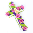 Against a clear, immaculate white background, a striking floral cross is presented at a gentle diagonal, the top left arm slightly raised so the arrangement feels alive and three-dimensional. The outline of the cross is meticulously formed with a plush band of white spray chrysanthemums, their many tight petals overlapping to create a soft, cushion-like border that defines the shape with precision. Just beyond this white halo, a delicate fold of pale green ribbon or leaf wrapping runs around the entire edge, echoing the fresh greens you might see in Oxford's Botanic Garden and giving the tribute a graceful, finished look. Within this frame, the cross bursts into colour and texture. Large, fully opened roses in vivid fuchsia pink and creamy pale yellow dominate the design, their spiralled petals appearing velvety and full of emotion. Between these showpiece blooms, clusters of lime-green button chrysanthemums add a playful, pom-pom effect, infusing the arrangement with energy and contemporary charm. Scattered among the roses and green mums are clusters of smaller, deep purple and violet flowers, likely statice or lisianthus, bringing a richer, more contemplative tone that balances the brightness. The harmony of pink, cream, green and purple makes the tribute feel both respectful and uplifting, like sunlight breaking through clouds over the spires of Oxford. Even, bright lighting ensures that each flower, from the tiniest floret along the border to the largest rose at the centre, is clearly defined. This floral cross would serve beautifully as a Christian funeral tribute or memorial arrangement for services in Oxford, expressing faith, gratitude and a vibrant celebration of a life well lived.