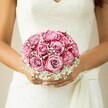 Held gently against an intricate white lace gown, this bridal bouquet embodies pure Oxford charm and timeless celebration. The arrangement catches the eye with its ensemble of two-tone pink roses-each bloom unfurling in perfect symmetry from a gentle, candlelit blush at the heart to a vivid magenta glow at the edges, reminiscent of the pastel sunsets over Port Meadow. Whispery white baby's breath floats between the roses, softening the look and adding an ethereal veil of texture, like the early mist that gathers along the Cherwell on summer mornings. The bride's lightly tanned hands, adorned with a barely-there, lustrous manicure, cradle the bouquet with pride and anticipation. Her sleeveless gown, rich with floral lace and a refined V-neckline, hints at both tradition and contemporary Oxford sophistication. A subtle sash detail trails at her side, echoing the understated elegance seen in wedding parties crossing the cobbled streets around Radcliffe Square. Set against a gently blurred, neutral background that seems to invite quiet reflection, the bouquet's hues and textures speak not just to the florist's artistry, but to the hope and joyful promise of the day. It's easy to imagine this bouquet being crafted on the High Street or delivered fresh that morning by a local specialist-making it a radiant, heartfelt focal point for weddings and romantic celebrations across Oxford.