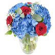 A lush, hand-arranged bouquet sits in a clear glass vase, the kind we favour in our Oxford studio, photographed against a clean white background so every blossom reads clearly. Dominating the composition are large, rounded clusters of bright, sky-blue hydrangeas; each cluster is a soft, cloud-like mass of small overlapping florets whose cool blue edges appear almost translucent in the bright light. Nestled through that billowy blue are three deep red roses, their velvety petals tightly spiralled and richly saturated, offering a dramatic, romantic punctuation to the arrangement. Sprays of tiny white baby's breath (Gypsophila) float between the larger blooms like flecks of early morning mist, lending delicate air and a subtle contrast that lightens the overall silhouette. Muted, rounded eucalyptus leaves and fresh green foliage weave around the stems, their silvery-green tones providing depth, gentle scent and an organic frame to the flowers. The clear vase has a gently rounded base and tapered top, and through it you can see the crisp green stems submerged in water, a visible sign of freshness and careful handling. Bright, natural lighting highlights the contrast of cool blue against warm crimson and the soft textures of petals and foliage, creating an optimistic, contemporary feel. This arrangement reads as both celebratory and intimate - equally suited to a sunlit table near the Botanic Garden, a thoughtful anniversary gift from a student returning to High Street, or a striking centerpiece that brings a sense of calm and joy to any Oxford home or small event.