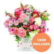 A generous, romantic bouquet overflows from a clear, square glass vase, captured against a pure white backdrop so each bloom appears crisp and luminous. The arrangement is dominated by roses in varying shades of pink, from the intense, almost jewel-like fuchsia and magenta tones to softer, pastel blush roses whose petals open in gentle spirals. Nestled among them are lush carnations in coral-pink, peach, and pale rose hues, their frilled petals adding a soft, billowing texture that makes the bouquet feel luxuriant and full. Threaded throughout are daisy-like chrysanthemums in lavender and lilac, each with a vivid green centre that injects a fresh, energetic note, balanced by touches of creamy white flowers that bring calm and clarity to the colour story. The blooms are supported and framed by abundant greenery: rounded eucalyptus leaves in a cool, silvery-green, richer dark green foliage, and occasional reddish-brown leaves that offer a hint of autumnal depth, reminiscent of the shifting tones along Oxford's Botanic Garden paths. The flowers sit securely in a transparent, rectangular vase whose lower portion is wrapped in a white band, printed with repeating red hearts and the loving inscription all my love, making the sentiment behind the gift unmistakable for anniversaries, Valentine's surprises, or heartfelt thank-yous. In the bottom right corner, an orange circular graphic displays the words VASE INCLUDED in bold white capitals, indicating that this bouquet arrives as a complete, ready-to-display gift. The overall impression is of a thoughtfully crafted arrangement, ideal for brightening living rooms in Summertown, cosy terraces off Iffley Road, or office spaces across central Oxford with a burst of colour and affection.