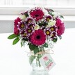 A vibrant mixed bouquet of fresh flowers stands proudly in a short, textured clear glass vase, centred against a softly glowing white background that feels like daylight streaming through an Oxford townhouse window. The arrangement is dominated by bold gerbera daisies in deep fuchsia and magenta, each with a dark, velvety centre that draws the eye immediately. Around them, clusters of bicolour chrysanthemums ripple through shades of rich purple at the petal tips, fading gently to crisp white with bright yellow centres, adding playful contrast and intricate texture. Creamy white spray roses, some fully open and others still in tight bud, are tucked between the larger blooms, lending a soft, romantic note and a hint of classic fragrance. Sprays of delicate white baby's breath weave through the display like tiny stars, creating a cloud-like lightness above the deep green foliage that forms the base. The glass vase is cinched at the neck with a simple bow of rustic twine, and from it hangs a small, rectangular light-coloured gift tag printed with a stylised pink rose motif and gentle script, suggesting a personal message without the writing being legible. The natural light highlights the freshness of each petal, making the colours appear luminous, as if the bouquet has just been arranged for a birthday, thank-you, or congratulations gift headed to a cosy home near Oxford's Covered Market or a college room off the High Street. The overall feeling is one of cheerful celebration and thoughtful care, a modern yet timeless floral design perfect for brightening everyday spaces or marking special moments with locally delivered flowers.