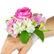 A refined wrist corsage sits gently on a light-to-medium skin toned left hand and forearm against a clean white backdrop, composed like a small wearable garden. At its centre is a large, fully opened deep fuchsia-pink rose, petals packed in a tight spiral that suggests a slow, fragrant unfurling; the rose reads as the confident focal point, glossy at the edges where soft studio light catches each curve. Surrounding it, clusters of soft blush-pink hydrangea florets add a cloud-like volume and delicate texture, their papery petals creating a cushioned base for the composition. Several pristine white alstroemeria (Peruvian lilies) fan out with four to six smooth petals apiece, each showing faint pale green streaking toward its throat, lending a crisp, luminous contrast to the warmer pinks. To the right, a small cluster of pale green alstroemeria buds and a few tighter pink buds promise more blooms, their downy calyxes and slender stems introducing a sense of gentle movement. Broad, emerald-green leaves cradle the entire arrangement, their rich sheen and firm vein structure grounding the palette and amplifying the colour warmth. The corsage is secured with an elegant multi-strand bracelet of small, lustrous white pearls that wraps around the wrist and trails partially along the back of the hand, offering classic sparkle against the skin. The overall impression is one of thoughtful craftsmanship-a sophisticated choice for a prom, a bridesmaid on her way to a ceremony at the Sheldonian Theatre, or anyone seeking a delicate, wearable accent that smells faintly of garden roses and spring greenery.