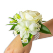 A delicate, hand-crafted white wrist corsage sits against a clean white backdrop, the arrangement extending from the bottom right of the frame toward the centre on a fair-skinned wrist. At its heart a creamy ivory rose unfurls in velvety layers, catching soft light along each petal's edge and giving the piece a warm, powdery glow. Below and slightly to the left a larger, trumpet-shaped white bloom-reminiscent of an alstroemeria or a small lily-opens to reveal a pale green-yellow throat that draws the eye inward and adds a whisper of spring freshness. Sprigs of tiny, cloud-like Gypsophila nestle between the main flowers, their airy white dots creating gentle texture against the rose's silky surface, while several bright green buds, still closed, promise more life and movement. Near the wrist a cluster of petite daisy-like white florets with sunny yellow centres offers a sweet, almost playful counterpoint to the formal rose. Broad, glossy deep-green leaves with a smooth, waxy sheen frame the composition, their cool tones setting off the pristine whites. The whole bouquet is fastened by a chic multi-strand pearl bracelet: numerous lustrous, round pearls sit close to the skin on an elastic band, their cool smoothness a tactile echo of the floral refinement. The arrangement suggests the hush of a church aisle or the soft music of a ballroom, perfect for prom, wedding vows, or a formal gala. As a local florist in Oxford might note, the scent is faint and fresh-like early morning in the Botanic Garden-evoking purity and quiet celebration without overwhelming the moment.
