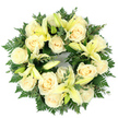 A quiet, dignified sympathy wreath viewed flat from above, composed of creamy ivory roses and soft pale yellow lilies set against a lush, green cradle of foliage - a composition that feels like a tender breath of consolation. About a dozen to fifteen fully opened roses, their petals spiralling inward with classic, velvety folds, form the wreath's warm, luminous core; pale yellow trumpet lilies are woven through the roses, some unfurled to display their graceful arched petals and subtle stamens, others kept in elegant bud form to add height and hopeful tension. The greens are deep and varied: feathery fern fronds give a lace-like edge, while rounded, matte leaves-reminiscent of eucalyptus-add cooler blue-greens and gentle weight. Soft, even daylight from above gives the cream tones a quiet glow against the clean white background, highlighting the wreath's dense, meticulously padded construction and its perfect circular silhouette, a timeless emblem of remembrance. There is a faint, comforting scent suggested in the arrangement-a powdery sweetness from the roses tempered by the green, almost citrus hint of the lilies-imagined through the layers of petals and leaves. The florist's hand is evident in the balanced spacing, the way buds and open blooms converse, and the thoughtful contrast between softness and structure. Suitable for funeral services, graveside tributes, or a solemn memorial, this wreath conveys peace, purity, and enduring affection. As a local Oxford offering, it echoes the gentle calm one finds in places such as Christ Church Meadow and the Botanic Garden, bringing a respectful, handcrafted presence to moments of farewell.