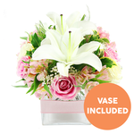 A romantic and uplifting bouquet is shown in a studio-style photograph, set against a bright white background that emphasizes the crisp colours and careful craftsmanship of the design. At the centre, elegant white Oriental lilies stretch open, their broad petals pristine and slightly curved, revealing tall, dark reddish-brown stamens and delicate pollen that give the flowers a dramatic focal point. Encircling the lilies is an abundance of soft pink roses, some tightly swirling in bud, others half-open, and a few fully blooming with layered, velvety petals, all combining to create a lush, rounded shape reminiscent of a hand-tied arrangement ready for an Oxford wedding or intimate dinner near the Covered Market. Between the roses and lilies, slender stems of pink and white alstroemeria peek through, their fine, speckled petals marked with subtle strokes of yellow that catch the light and invite a closer look. Light clusters of white baby's breath float throughout the arrangement, softening transitions between blooms and giving a cloud-like halo to the overall design. The flowers are surrounded by rich greenery, including broad, glossy leaves that anchor the composition and finer, feathery foliage that introduces gentle movement, like a breeze through the gardens along the Oxford canal. All stems are gathered into a contemporary square clear glass vase, the transparency revealing the neat structure below the waterline. Around the vase, a pale pink satin ribbon is wrapped and tied, adding a quiet, polished detail that makes the piece feel perfectly gift-ready. In the bottom right corner, a striking circular orange badge bears the words VASE INCLUDED in bold white capitals, clearly indicating that the container is part of what is shown. The overall impression is one of tenderness, freshness, and thoughtful design, ideal for same-day flower delivery in Oxford for birthdays, anniversaries, or simply to brighten someone's home.